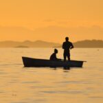 Two people fishing on a calm lake at sunset, silhouetted by the golden sky.